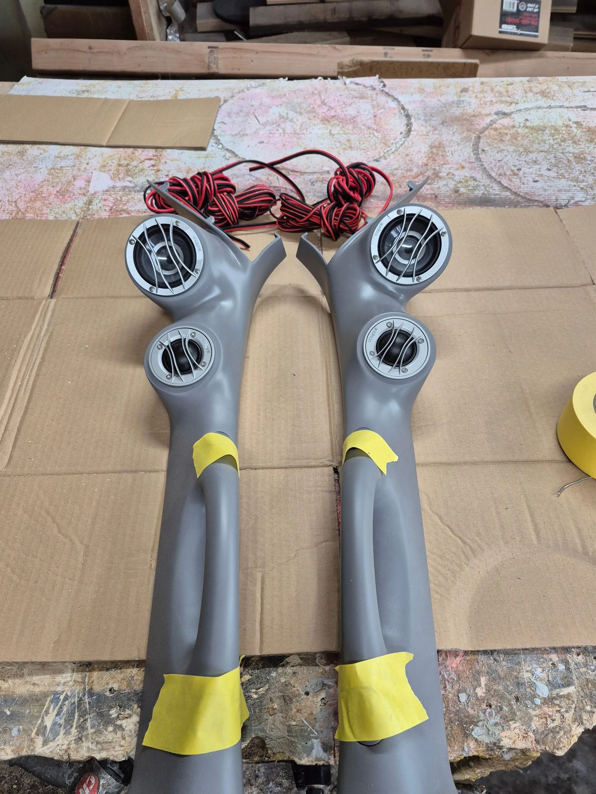 Pair of gray car pillar speaker mounts with speakers installed, masking tape, and speaker wire.