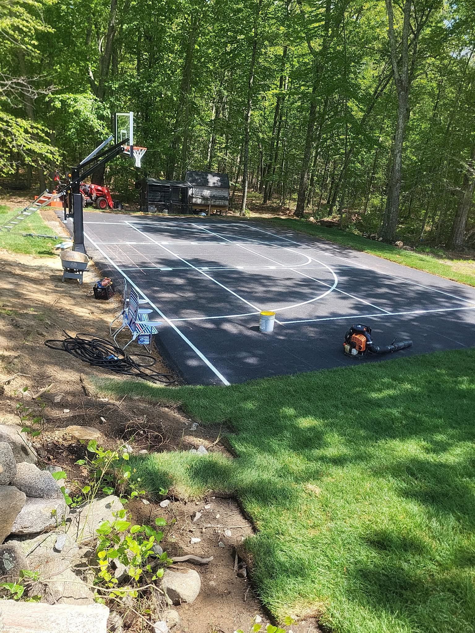 Outdoor basketball court under construction, with fresh black asphalt, white markings, and trees surrounding it.