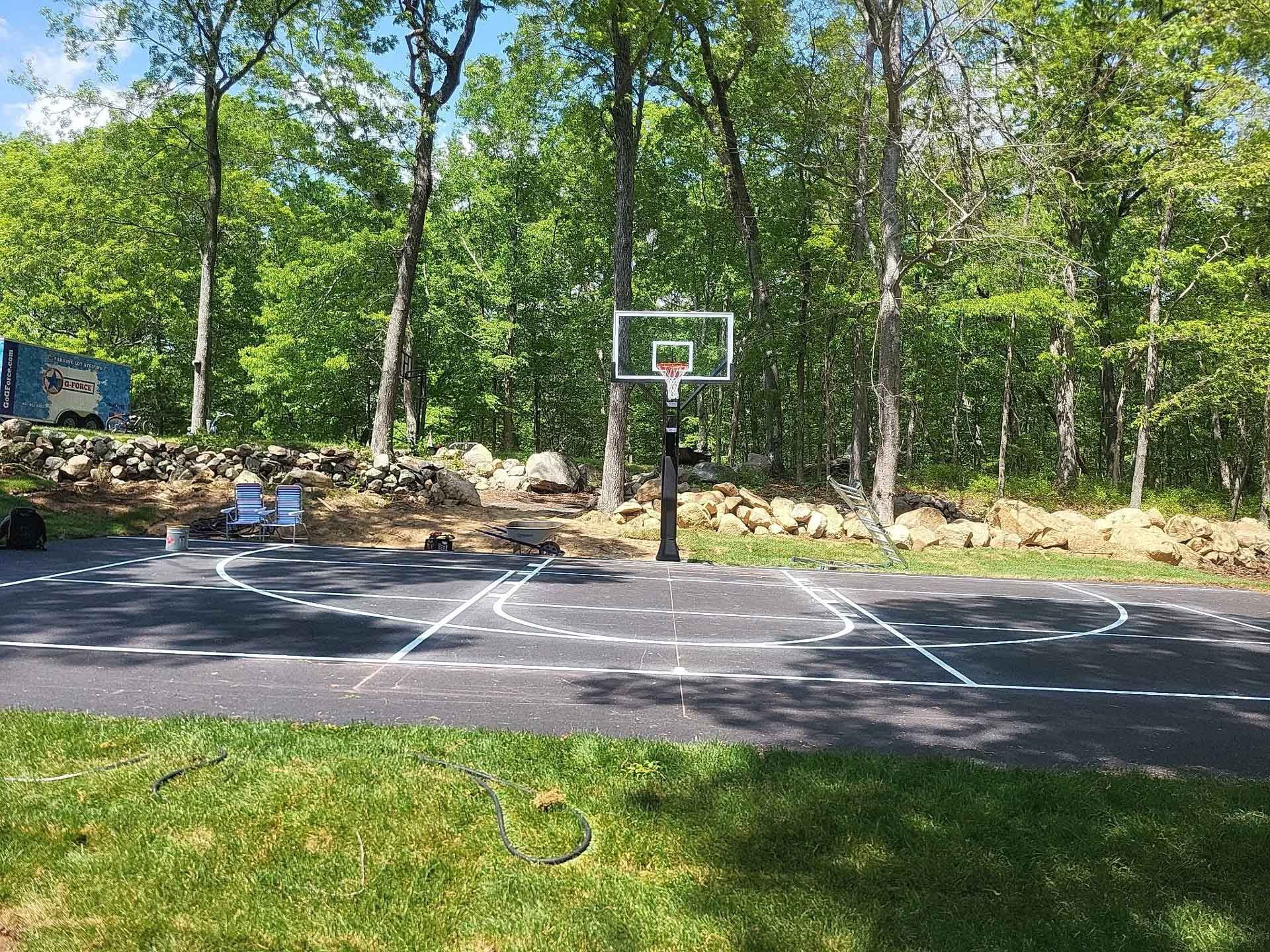 Outdoor basketball court in a wooded park, with trees, painted lines, and a hoop near a stone wall.