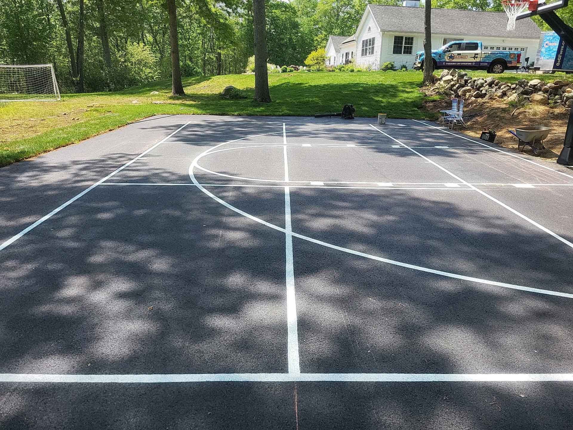 Empty asphalt court with white painted lines beside a wooded yard and house.