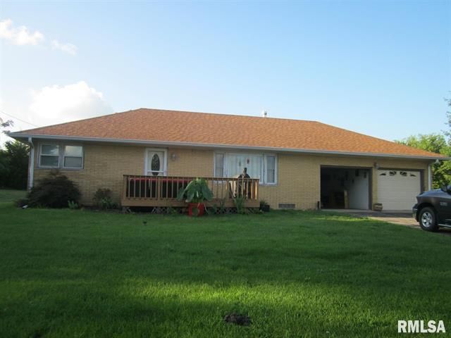 A ranch-style house with a tan exterior, brown roof, small deck, and attached garage, on a grassy lawn.
