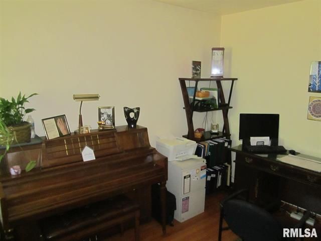 A room with a piano, desk, and shelving unit against light-colored walls.