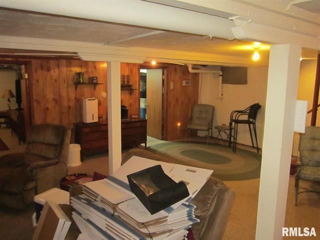 Basement with wood paneling, furniture, and clutter. Brightly lit, with support beams.