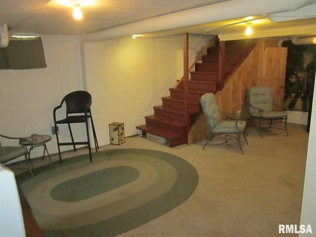 Basement room with stairs, chairs, rug, and overhead lighting. The walls are white and wood.