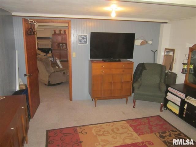 Bedroom with TV on a wooden dresser, an armchair, and a doorway to a cluttered storage space.