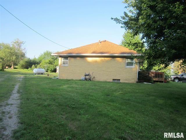 Yellow brick house with brown roof, situated in a grassy yard; trees and a propane tank are visible.