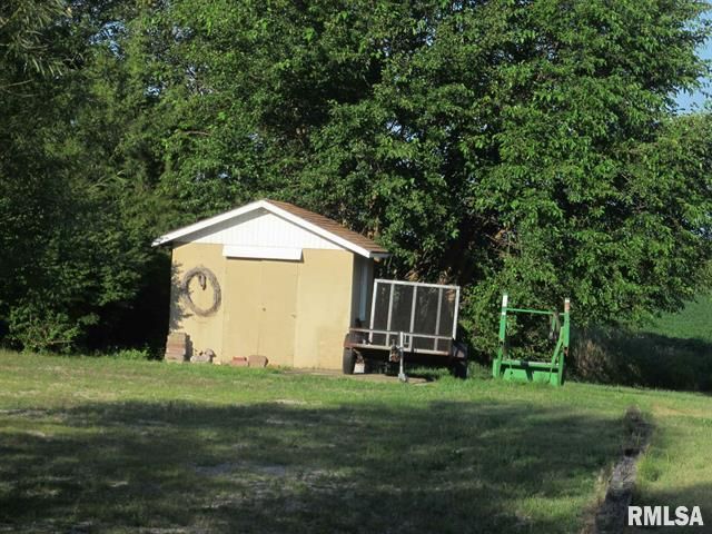 Small, tan shed with white roof, a trailer, and green metal equipment next to a tree in a grassy yard.