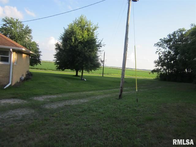 Yellow house with green yard, tree, utility pole, and fields in the background under a blue sky.
