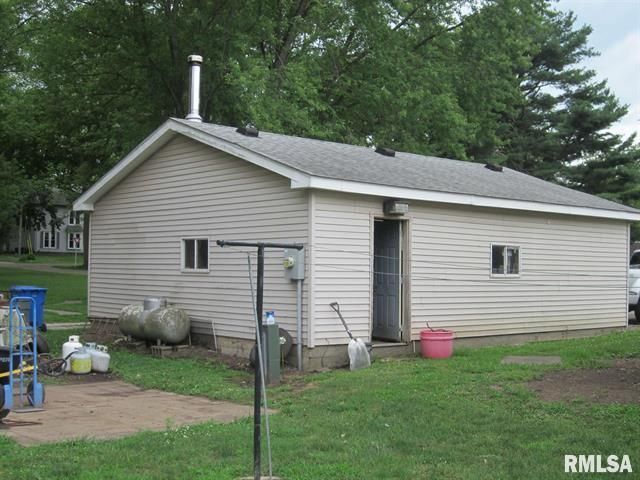 A tan-sided outbuilding with a chimney, door, windows, and propane tanks, in a grassy yard