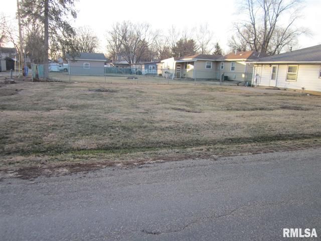 Empty, grassy lot in a residential area. Several houses and trees are visible in the background.