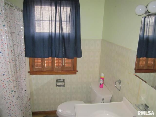 Bathroom with toilet, sink, window with blue curtain and brown shutters, and light green walls.