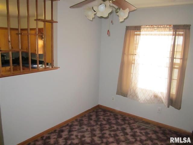 An empty room with a brown floral carpet, window with tan curtains, and a kitchen pass-through.