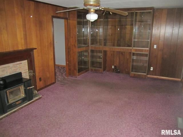 Living room with dark wood paneling, purple carpet, built-in shelving, fireplace, and a ceiling fan.