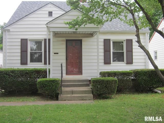 Small white house with gray shutters, front steps, and a red door behind trimmed hedges.