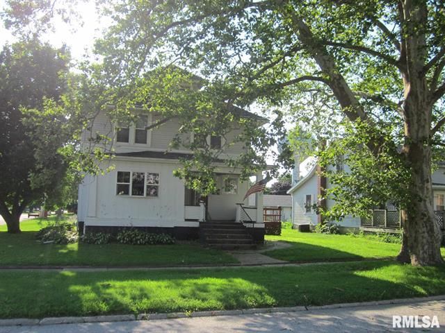 Two-story white house with front porch, shaded by large trees on a sunny lawn