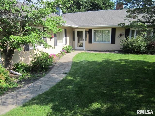 Single-story beige house with a curved stone walkway and green lawn under trees