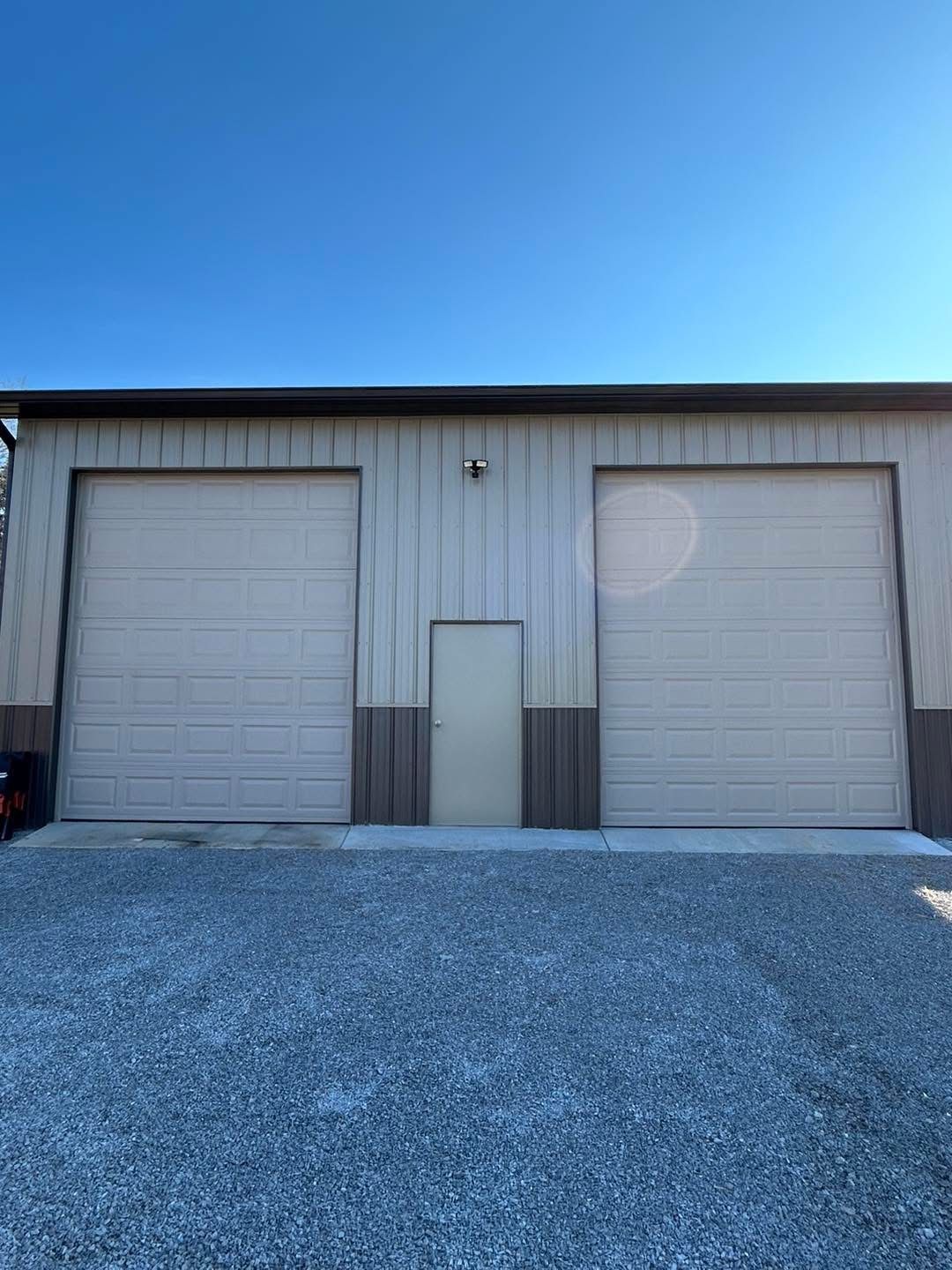 Exterior view of a light-colored metal building with two garage doors and a small door in the center; the ground is covered in gravel.