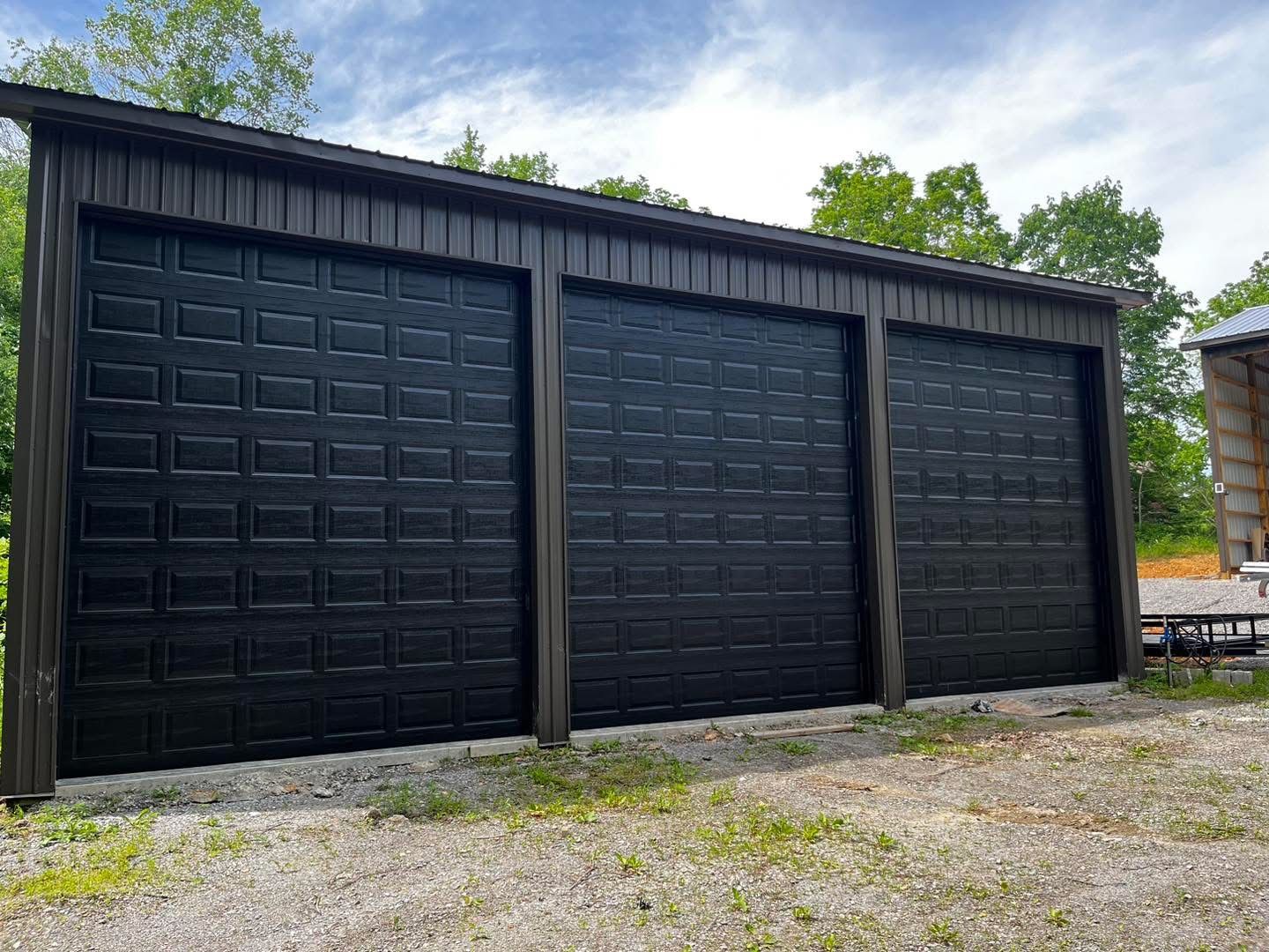 Three-bay garage with black doors and dark brown trim, set in a gravel lot under a partly cloudy sky.
