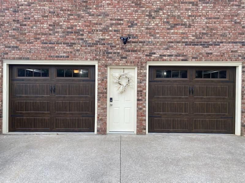 Two brown garage doors flank a cream-colored entry door, set against a red brick wall. A concrete driveway is in front.