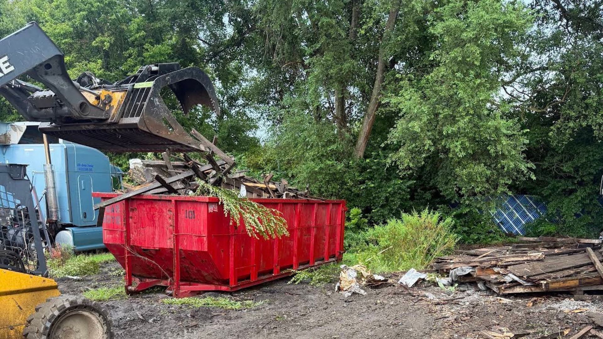 Excavator dropping brush into a red dumpster at a muddy outdoor worksite beside trees and debris