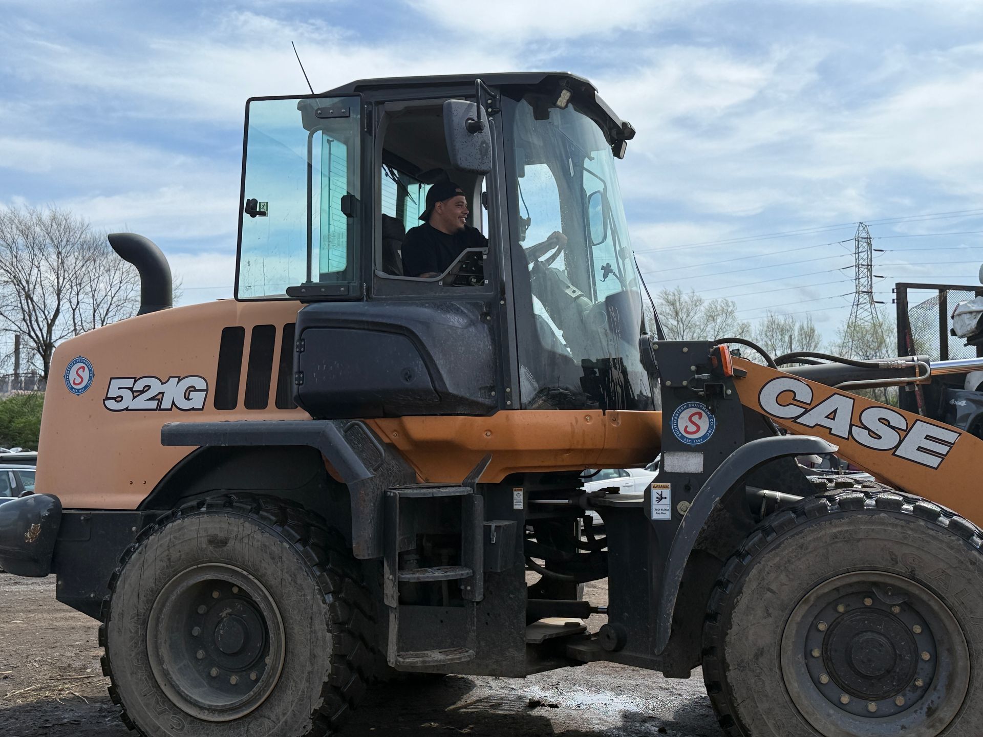 A person operates a tan Case 521G wheel loader on a gravel surface under a cloudy sky.