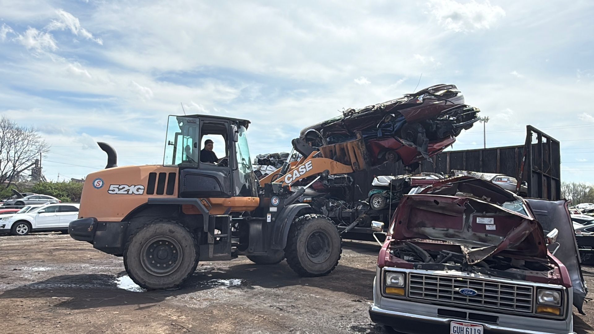 A Case loader moves a load of scrap metal into a truck container at a salvage yard, with a crushed car in the foreground.