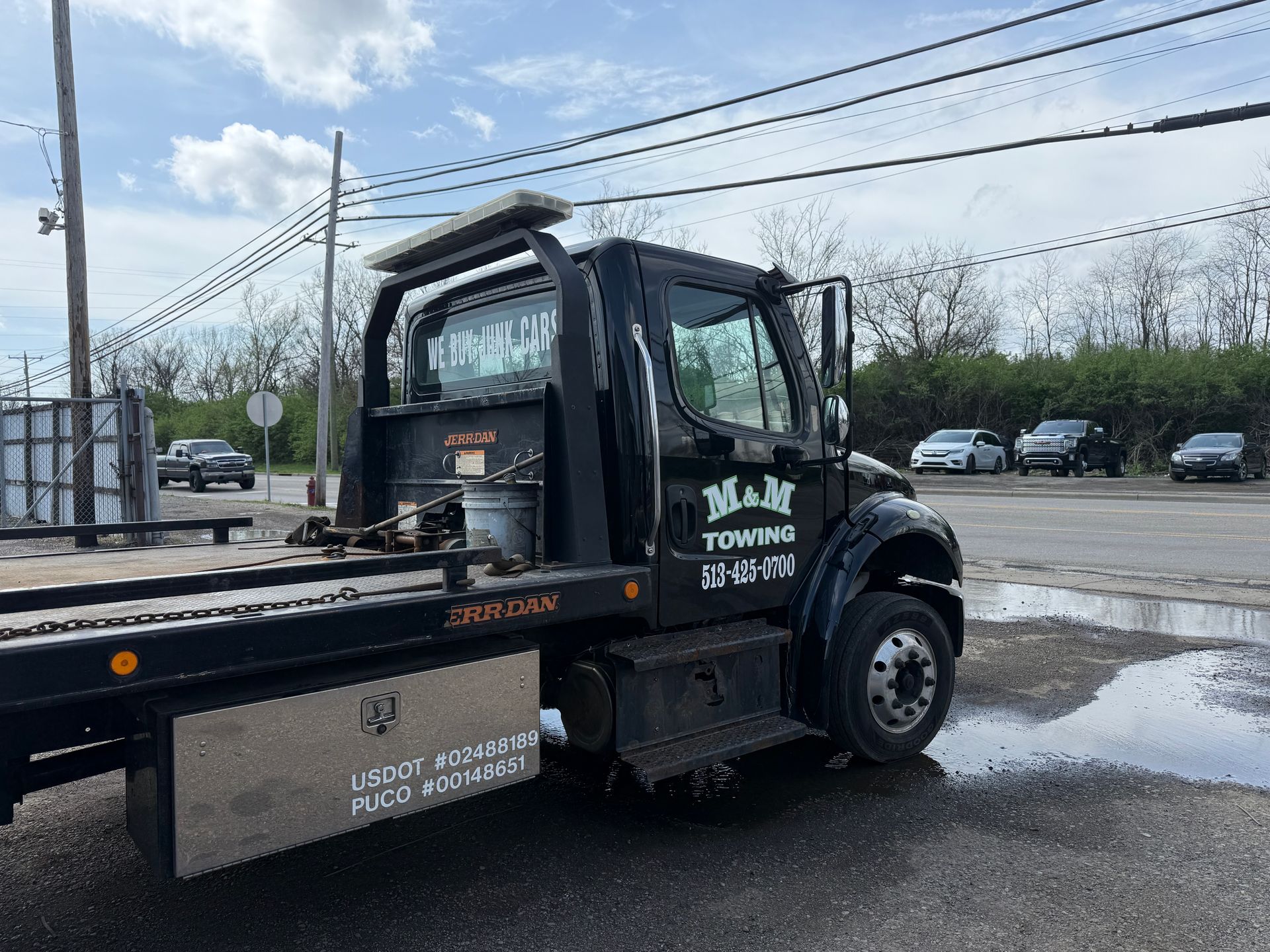 A black M&M Towing flatbed truck parked on a gravel lot under a cloudy sky.