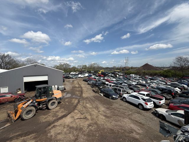 Wide outdoor car lot with rows of parked vehicles, a warehouse, and a front loader under a partly cloudy sky