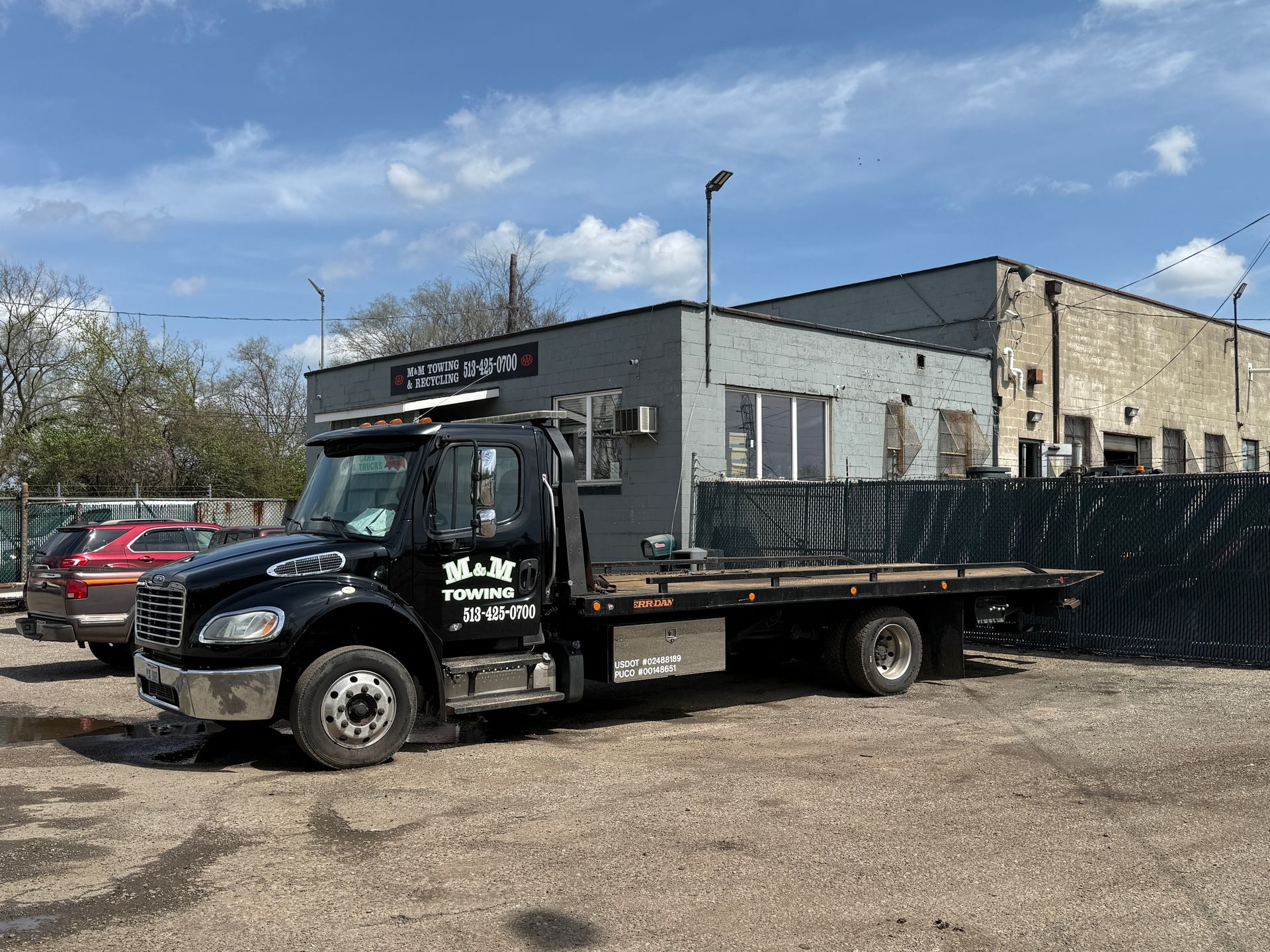 A black flatbed tow truck parked on a gravel lot in front of a grey industrial building under a blue sky.