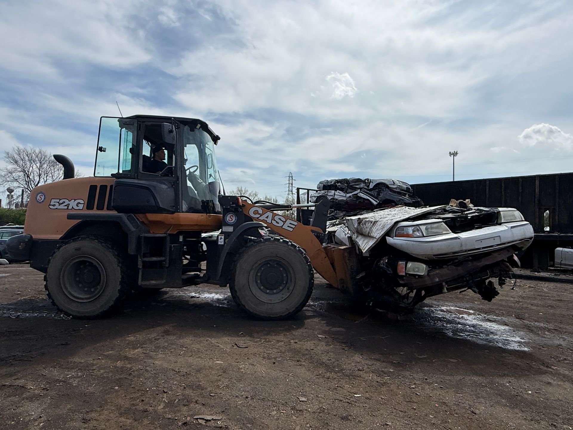 A tan CASE wheel loader moves a crushed white car in a scrap yard under a cloudy sky.