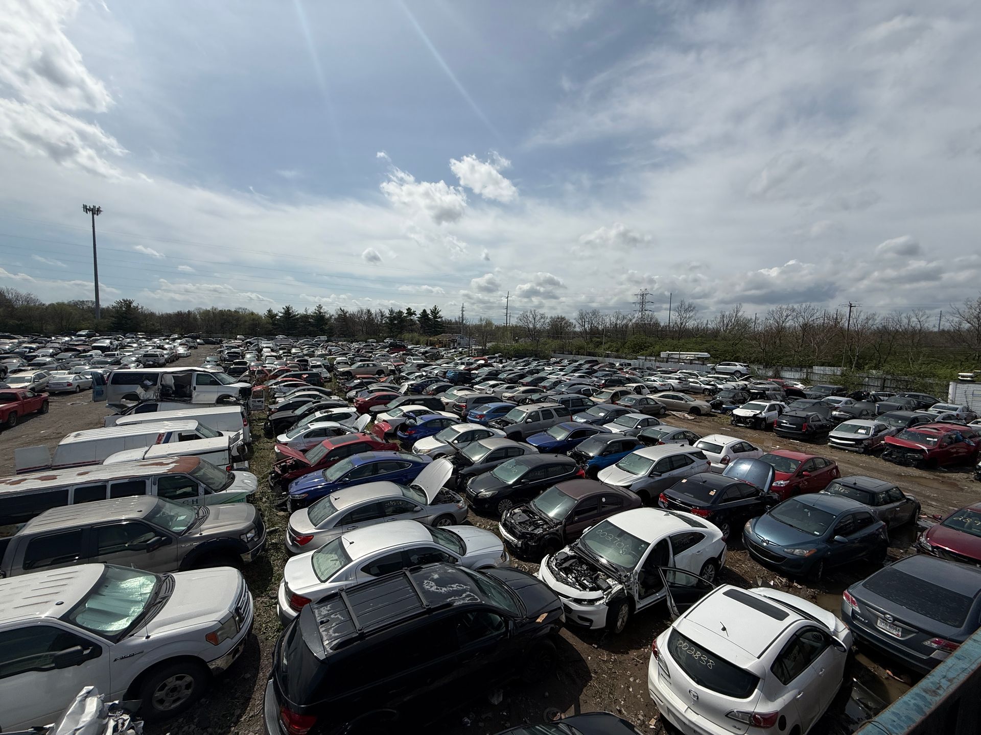 An expansive salvage yard filled with many damaged and parked vehicles under a bright, cloudy sky.