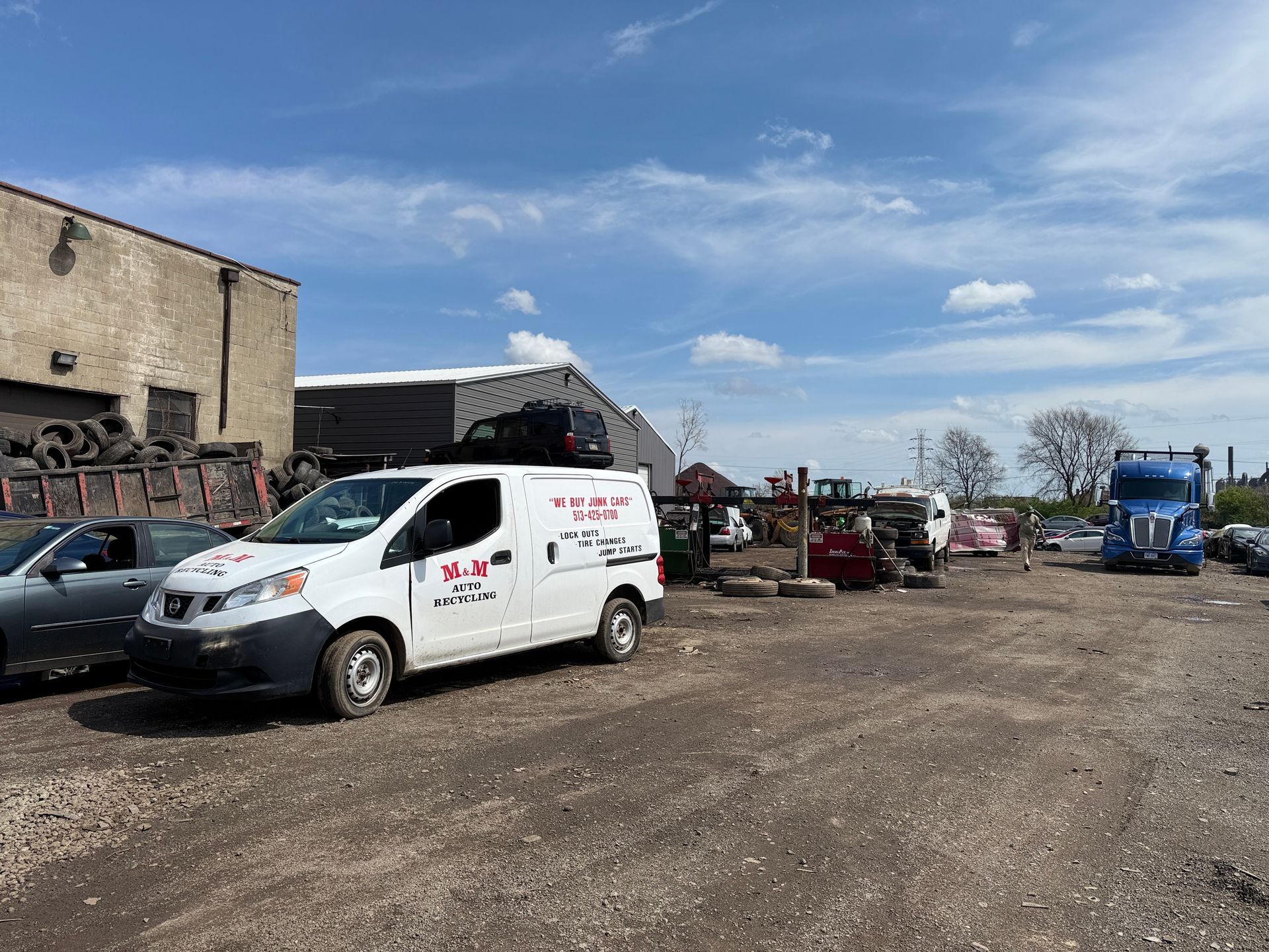 A white van parked in an outdoor salvage yard with various vehicles and scrap metal under a blue, cloudy sky.