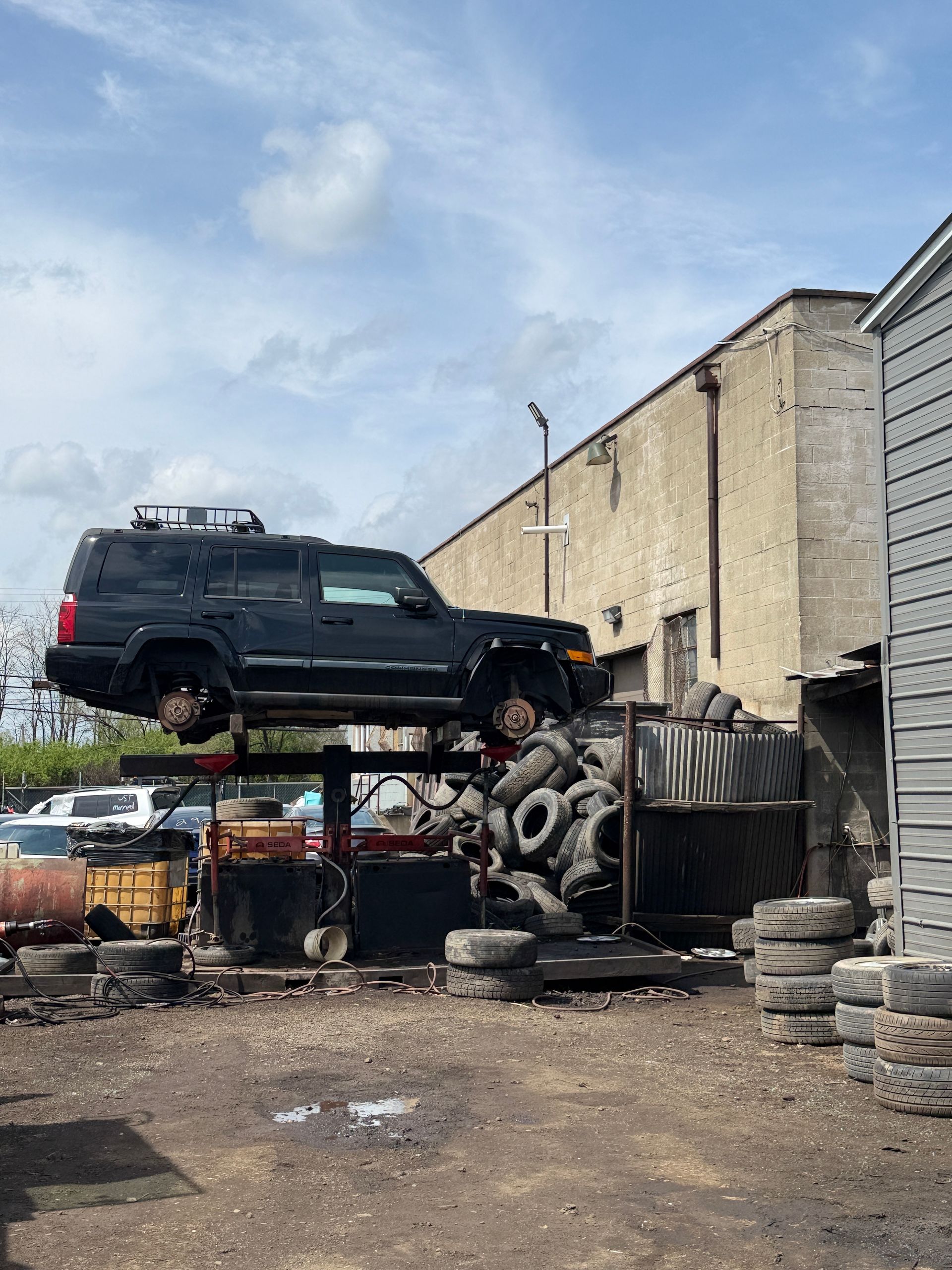 A black SUV sits on an elevated metal rack in an outdoor salvage yard surrounded by piles of discarded tires.