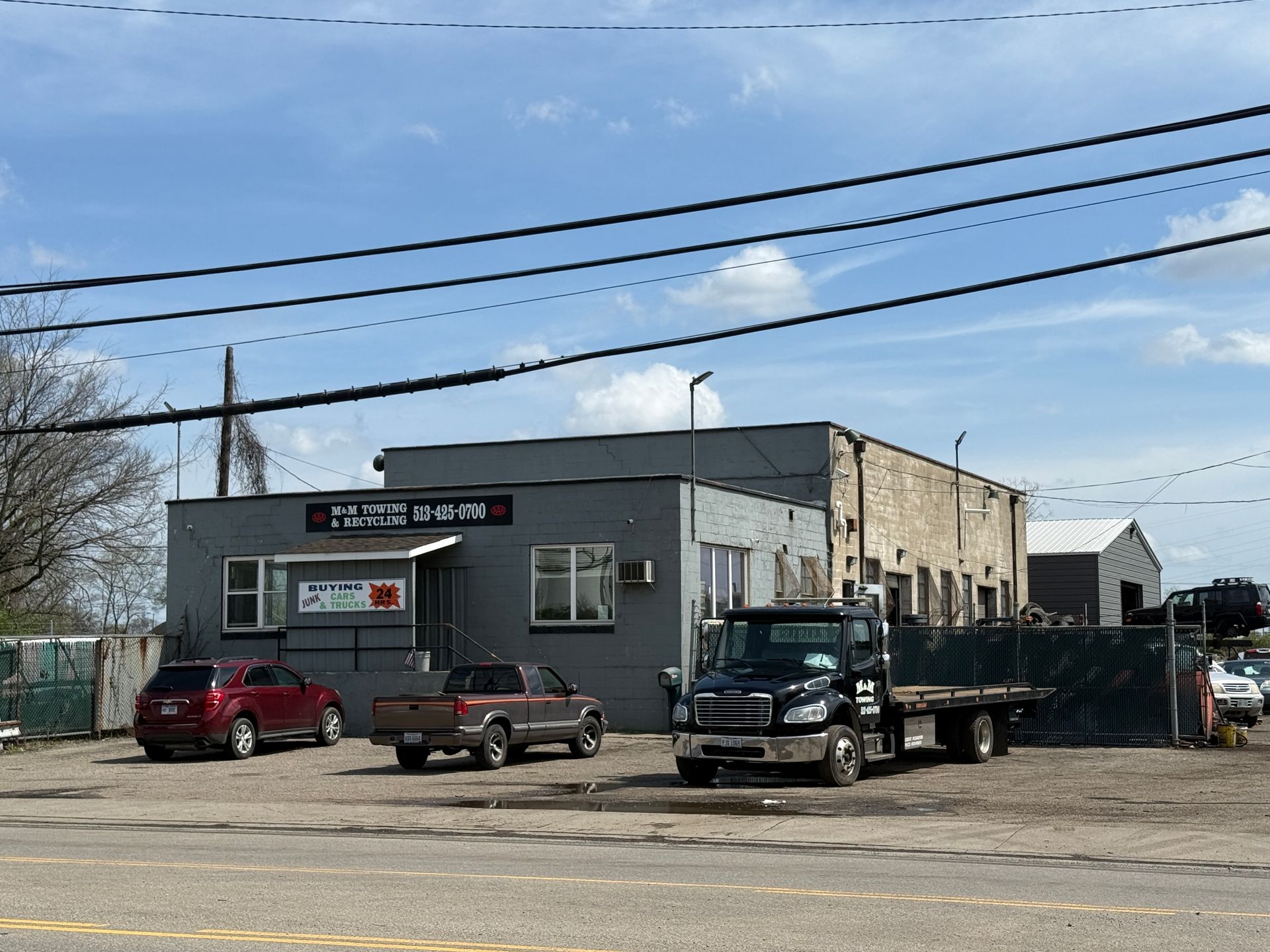 A grey commercial building with a dark-colored SUV, a pickup truck, and a large flatbed truck parked in a gravel lot.