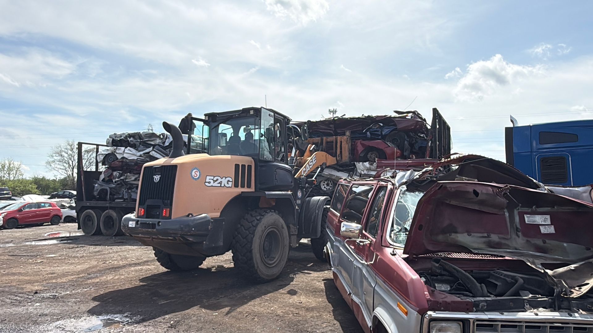 An orange front-end loader moves scrap vehicles at a junkyard under a bright, cloudy sky.