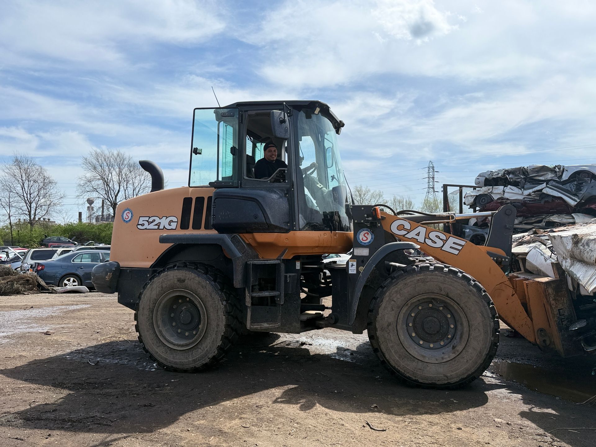 A Case 521G wheel loader parked in a junkyard with cars visible in the background under a blue, cloudy sky.