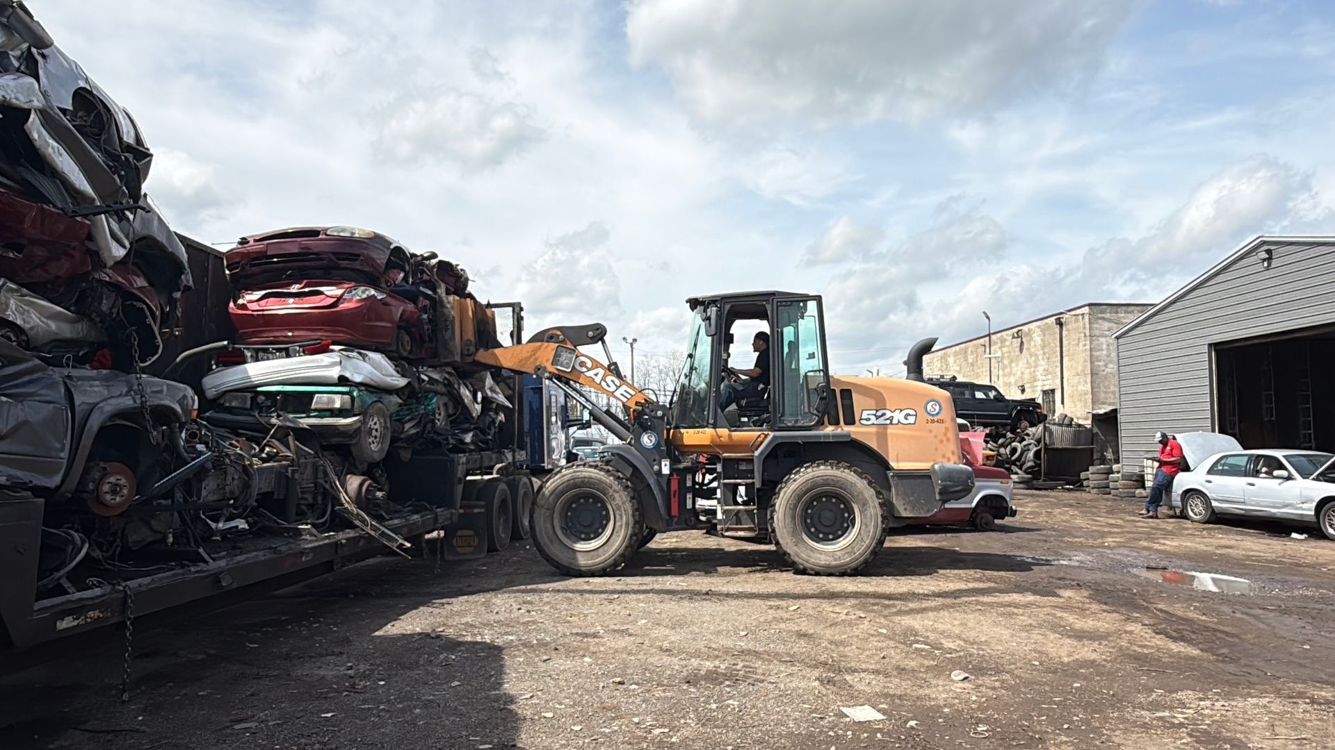 A construction vehicle lifts a stack of salvaged cars at an outdoor scrap yard under a cloudy sky.