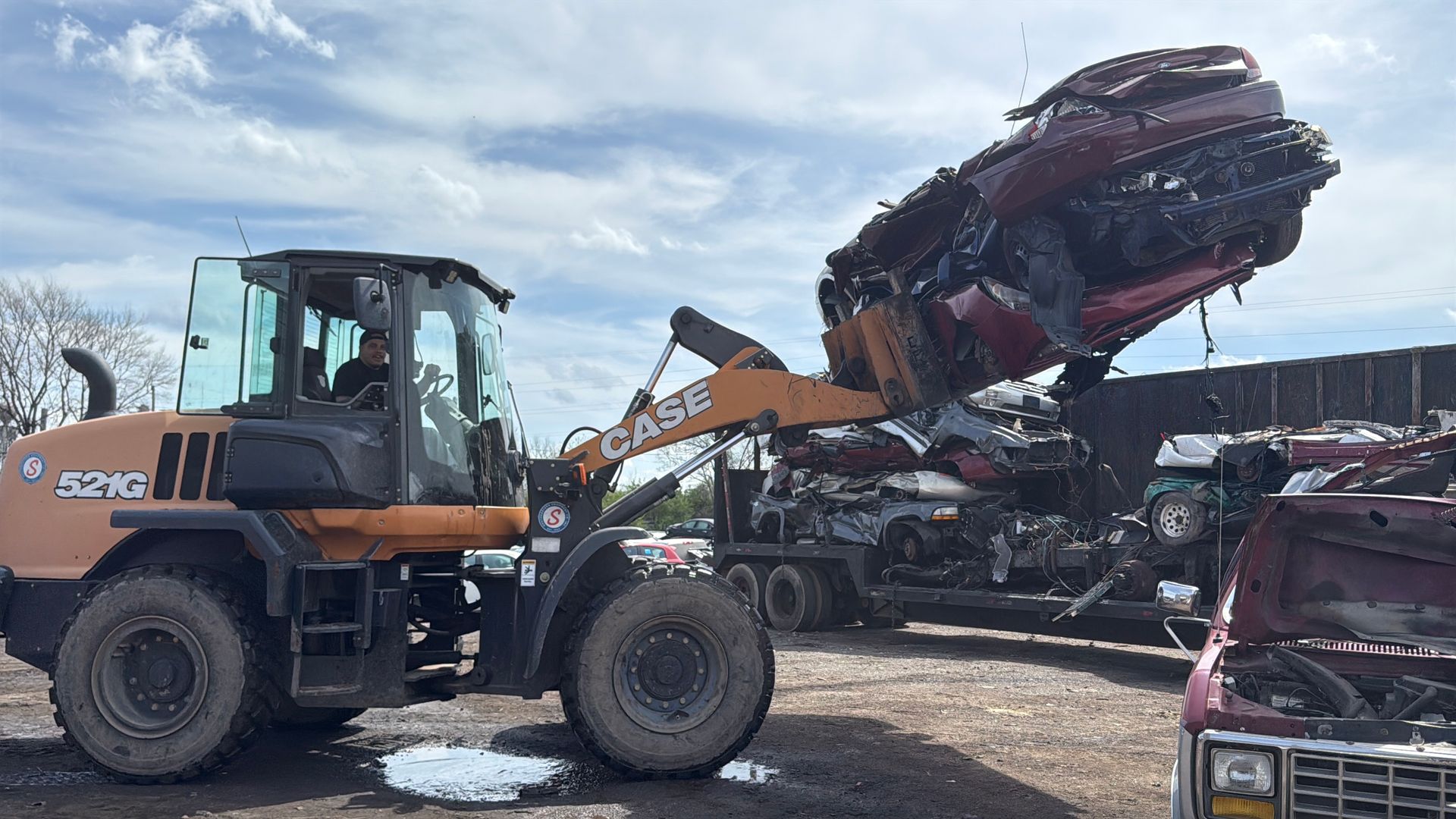 A yellow CASE wheel loader lifts a crushed maroon vehicle into a large metal container at a scrapyard under a cloudy sky.