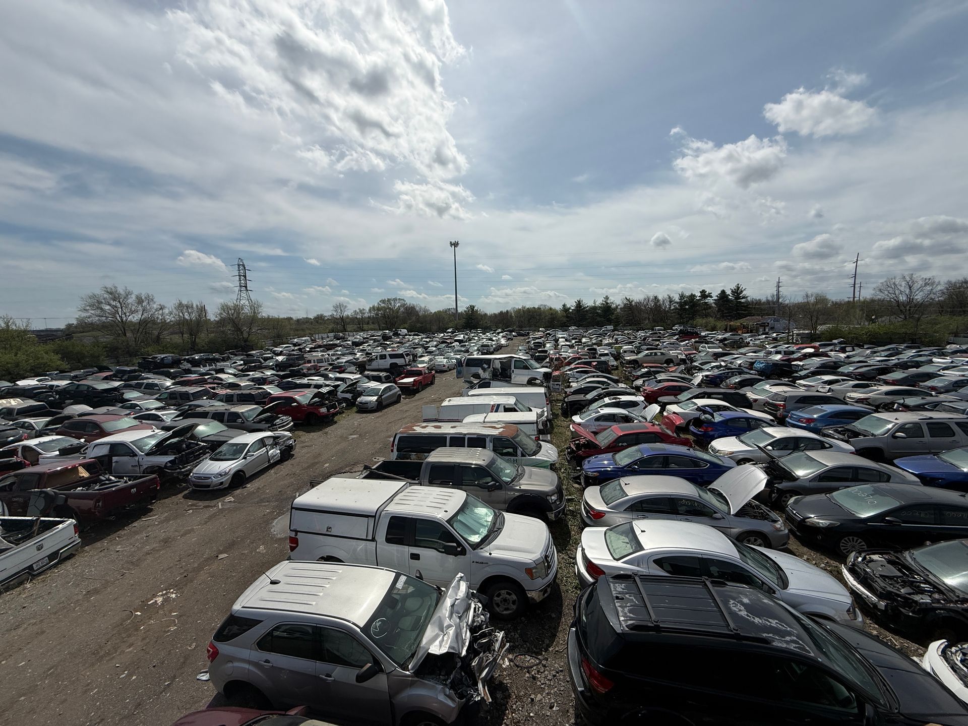 A wide-angle view of a vast salvage yard filled with many parked, damaged, and salvaged vehicles under a cloudy sky.