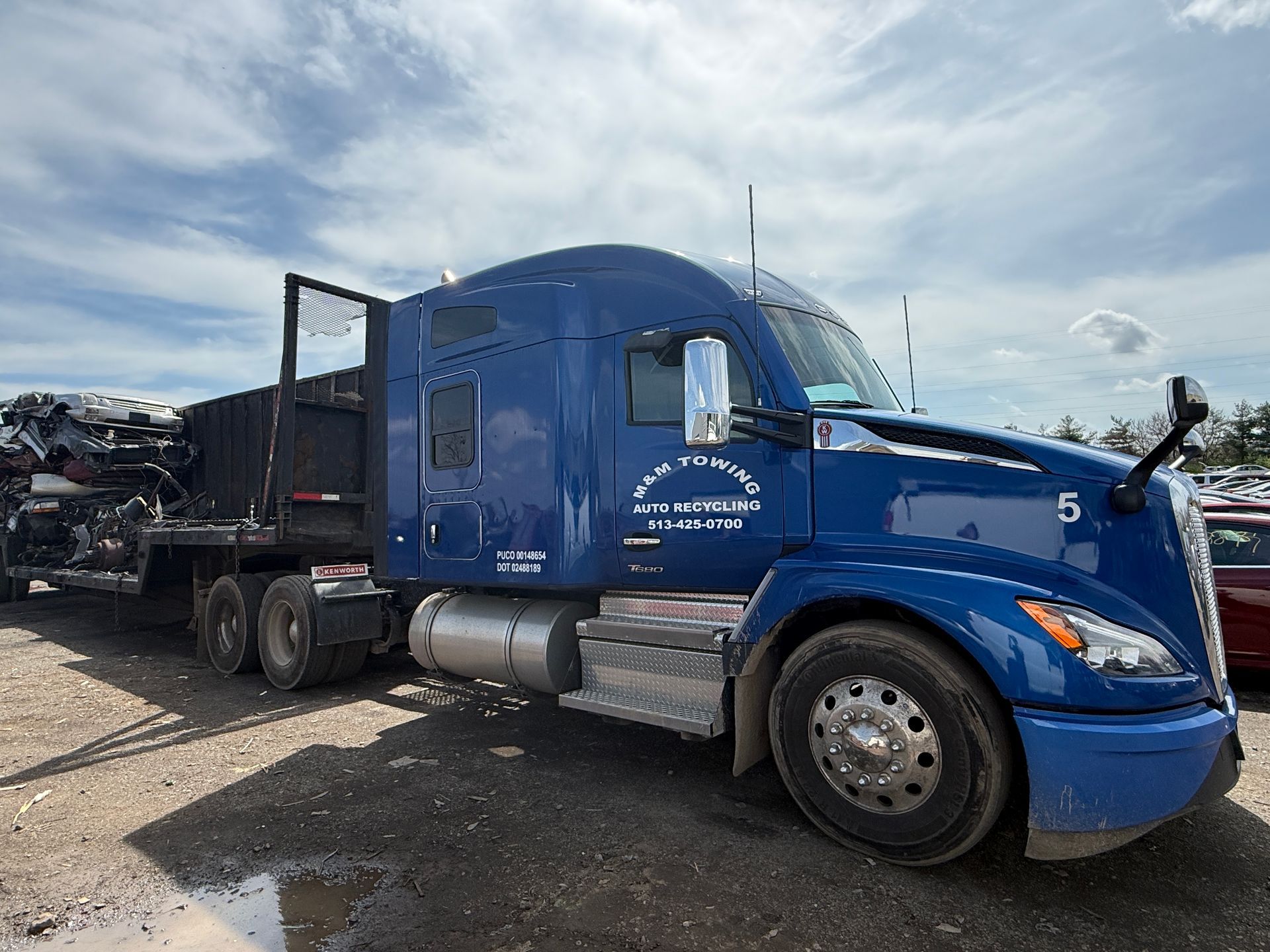 A blue semi-truck with a flatbed trailer carrying crushed cars is parked on a dirt lot under a cloudy sky.