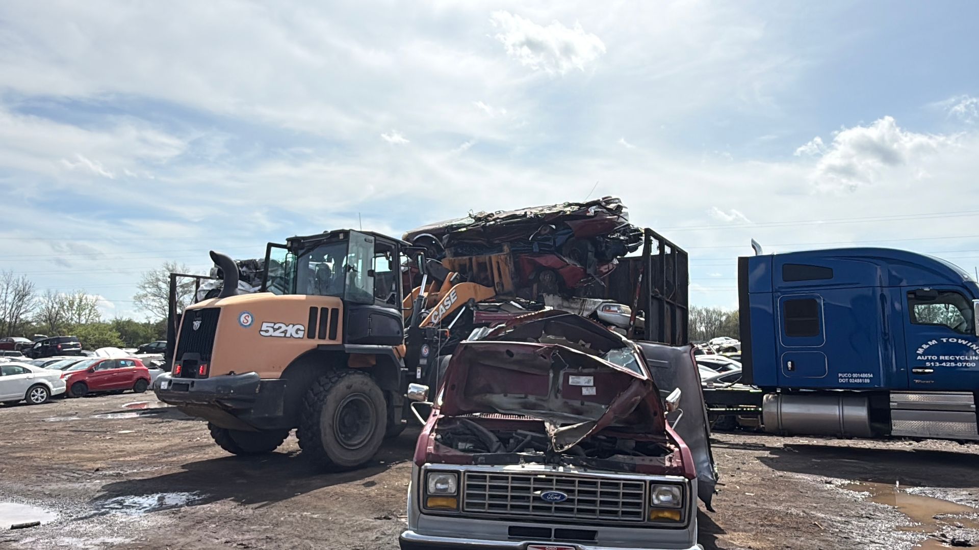 A bright orange loader works in a junkyard, loading salvaged scrap metal onto a truck near a blue semi-truck cab.