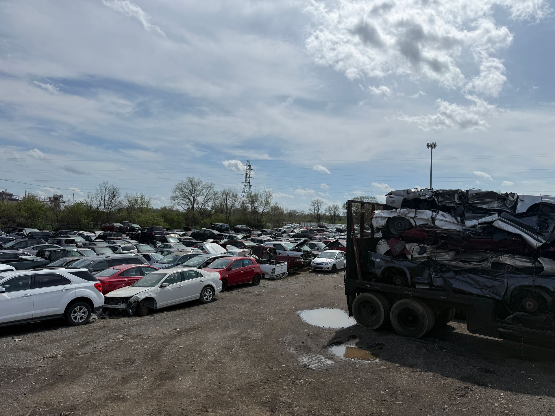 A salvage yard filled with many parked, damaged cars and a truck loaded with a stack of flattened vehicles.