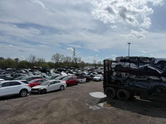 Large car lot with rows of vehicles under a cloudy sky, and a loaded truck at right.