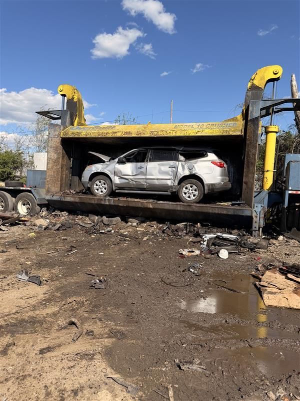 A silver SUV sits inside a large, industrial yellow car crusher on a muddy lot under a clear blue sky.