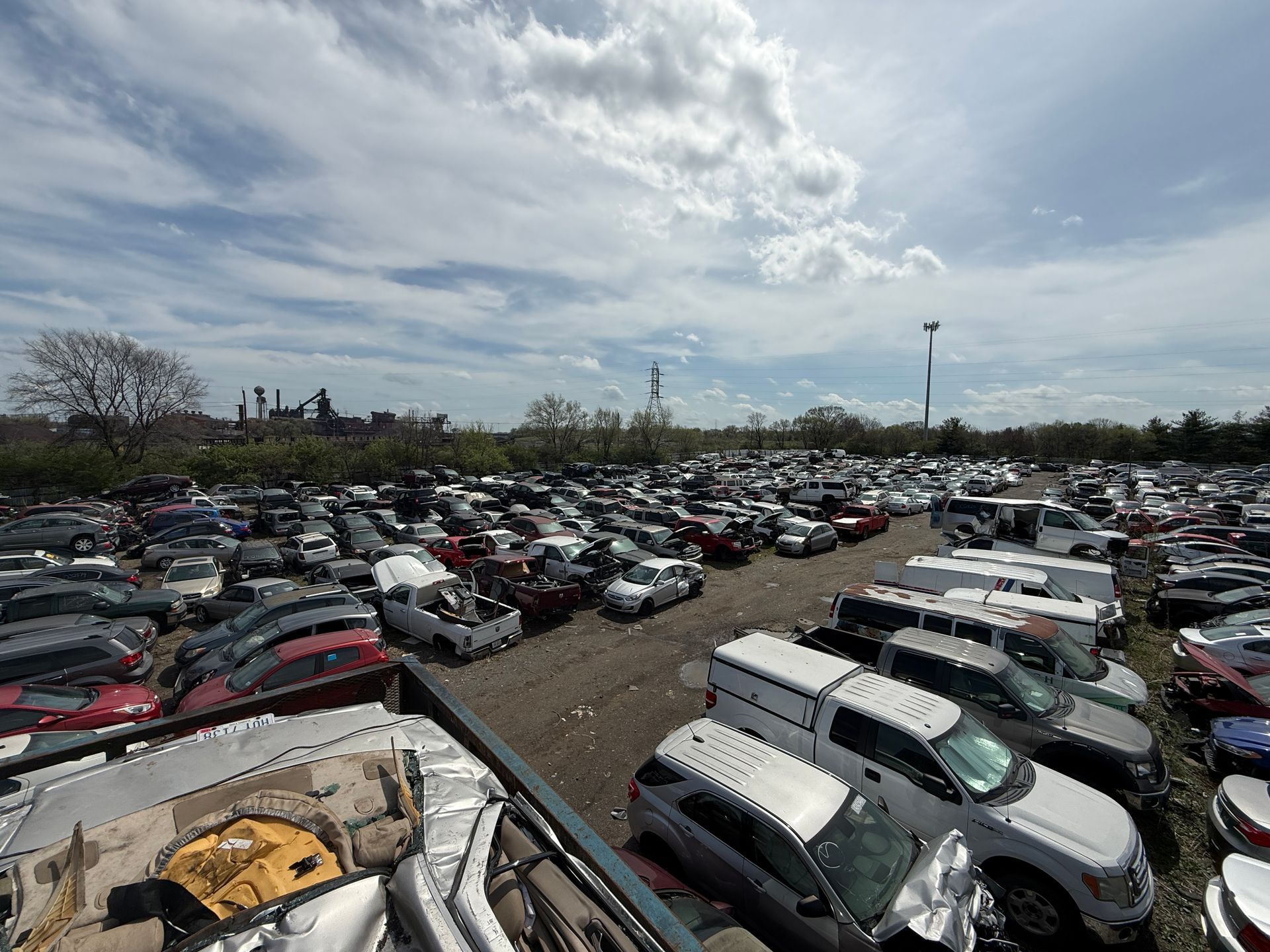 A wide-angle, slightly elevated view of a crowded junkyard filled with many wrecked cars under a bright, cloudy sky.