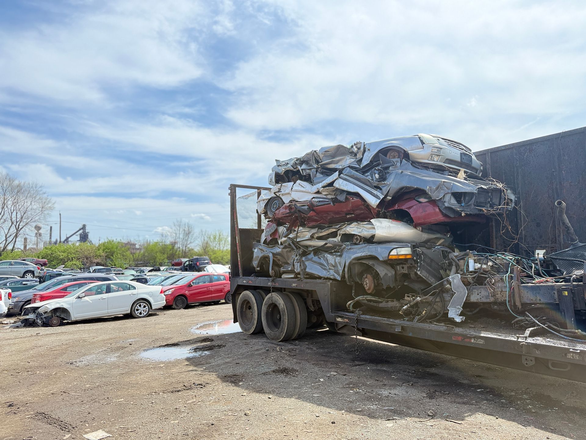 A trailer piled high with crushed cars at a salvage yard, with other vehicles parked on the gravel lot nearby.
