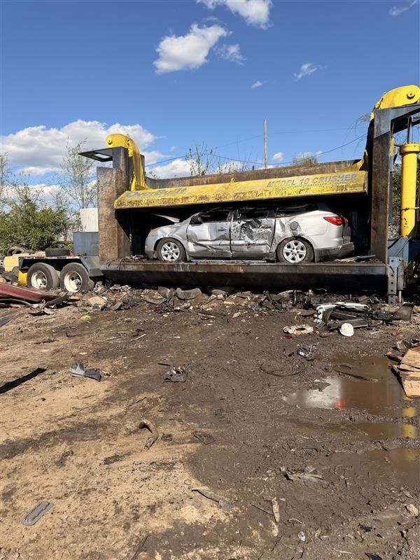 A damaged silver car sits inside a yellow hydraulic car crusher in an outdoor, dirt-covered junkyard under a blue sky.