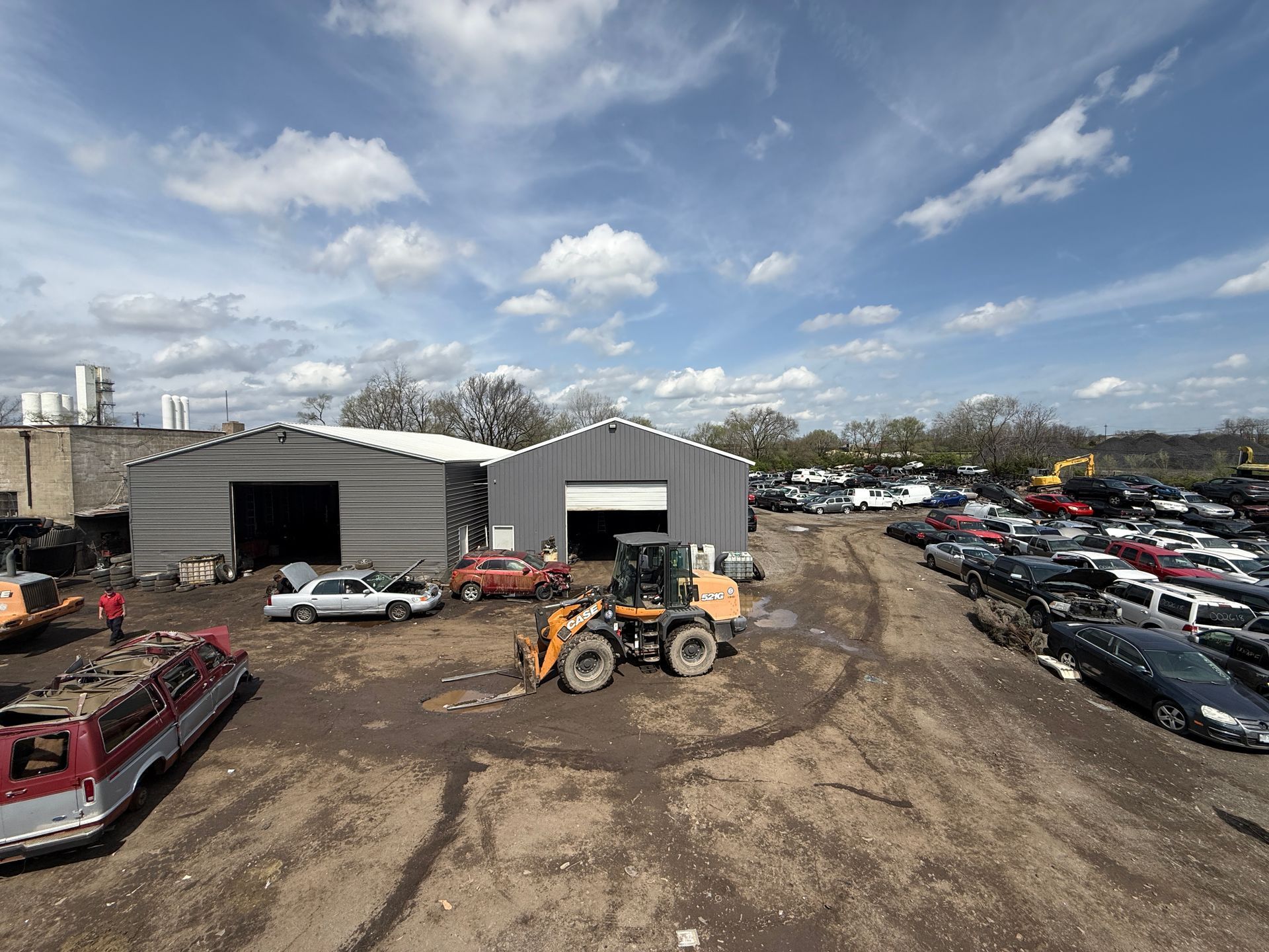 An aerial view of a salvage yard with gray warehouse buildings, a yellow loader, and rows of parked cars under a blue sky.