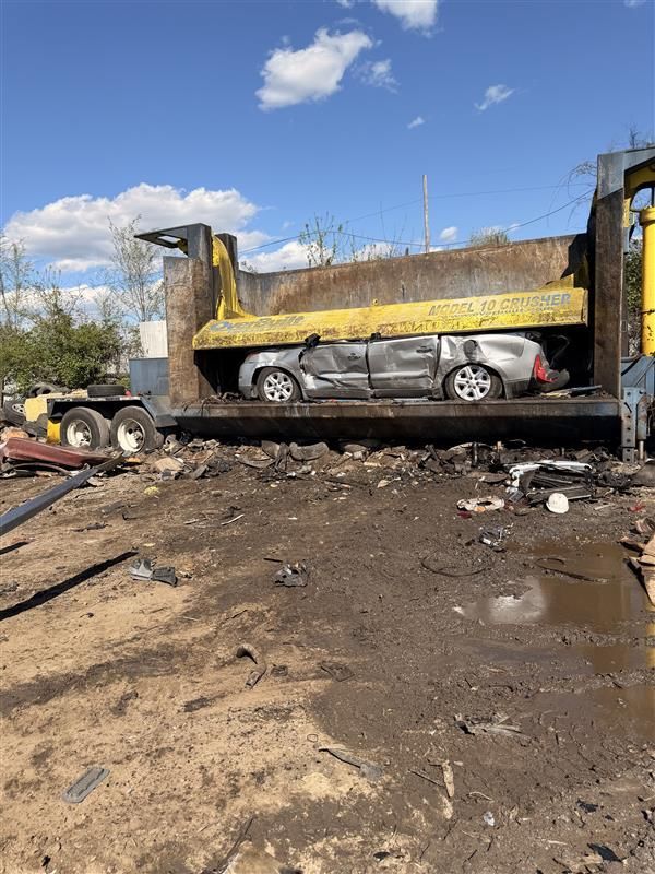A crushed silver car sits inside the metal bed of a yellow industrial vehicle at a scrapyard under a blue, cloudy sky.