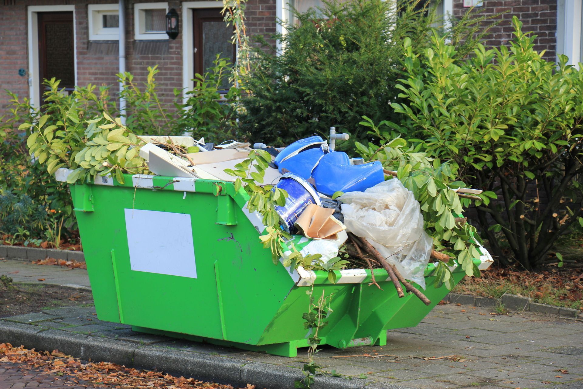 Green dumpster overflowing with trash and yard waste on a sidewalk outside a house